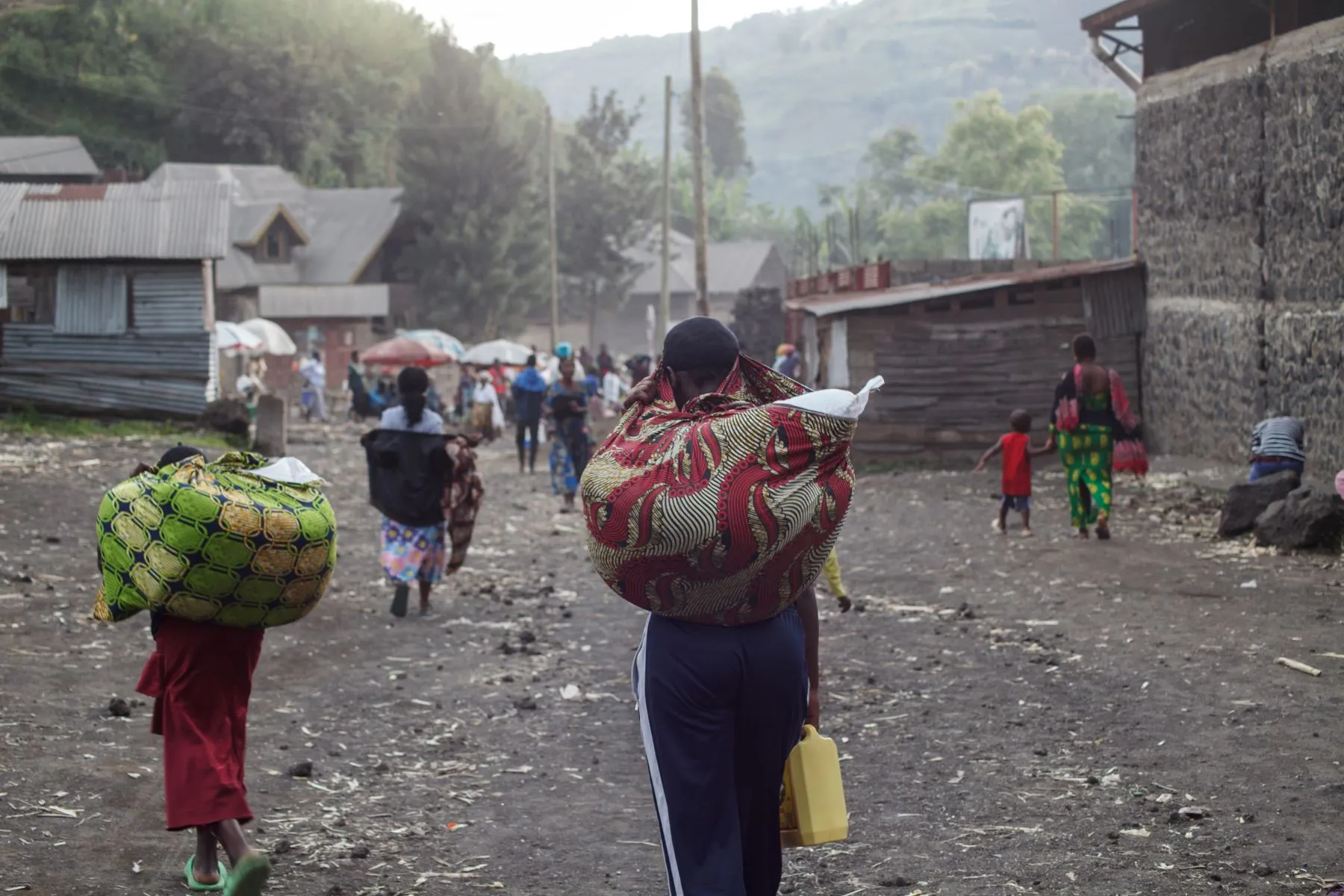 A group of people seen from behind, walking while carrying belongings wrapped in cloth on their backs.