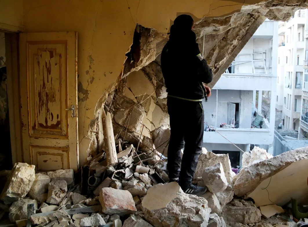 Rubble-filled room with a large hole in the wall; woman looks out at ruined buildings
