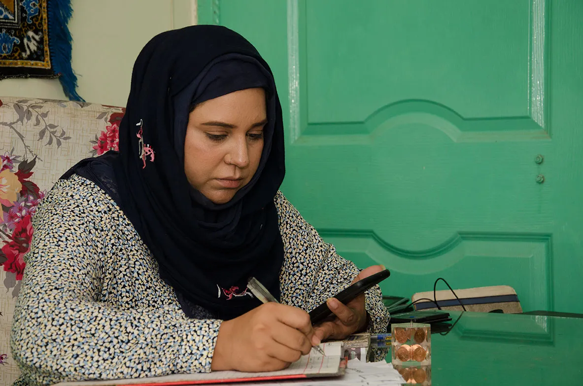 A Pakistani woman wearing a black head scarf sits in a chair. She is looking at her phone while copying down information in a notepad.