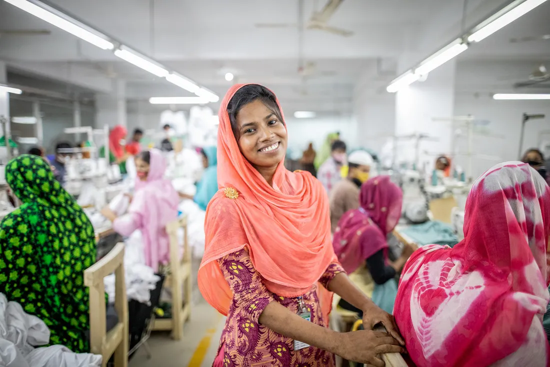 A woman wearing bright pink smiles at the camera. Behind her sit many women wearing brightly colored clothing and working at their stations.