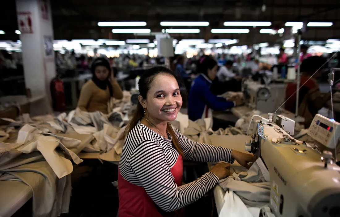 A woman working at a sewing machine looks up from her work. Behind her, many other women work at their stations.