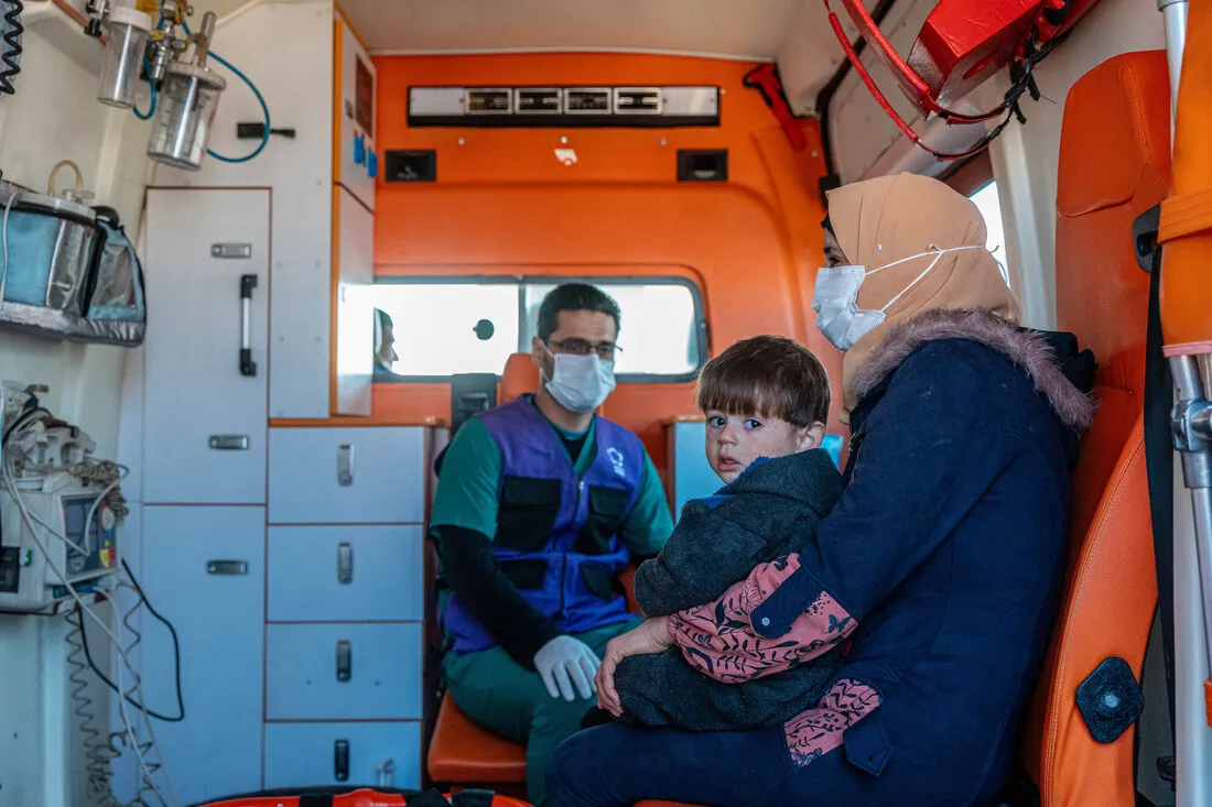 A mother in hijab and a facemask holds her child in the back of a medical transport vehicle on their way to the Al-Amal hospital.